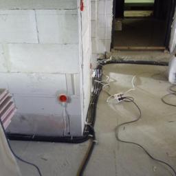 AMW Projekt - Surowy stan: Instalacja elektryczna w trakcie montażu w budynku z bloczków betonowych; widoczne kable w peszlach mocowane do ściany, puszki instalacyjne w kolorze pomarańczowym i przedłużacz...