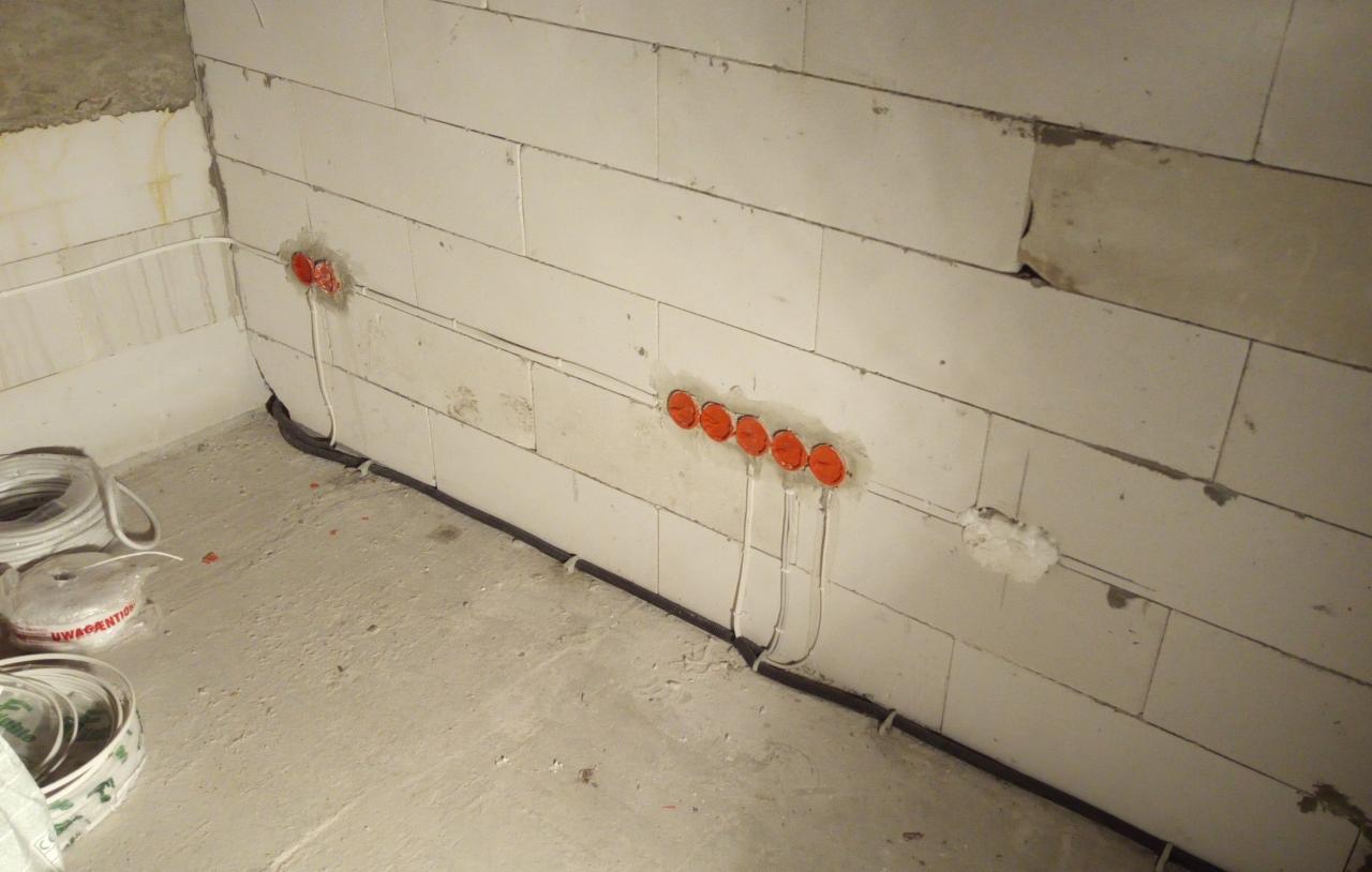 Ściana z betonu komórkowego z zamontowanymi puszkami elektrycznymi z pomarańczowymi pokrywkami, prowadzone kable w peszlu, widoczne zwoje kabli w tle.