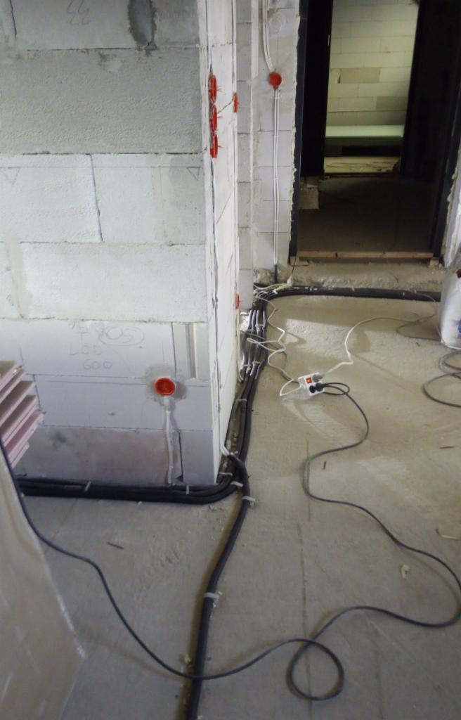 Surowy stan: Instalacja elektryczna w trakcie montażu w budynku z bloczków betonowych; widoczne kable w peszlach mocowane do ściany, puszki instalacyjne w kolorze pomarańczowym i przedłużacz...