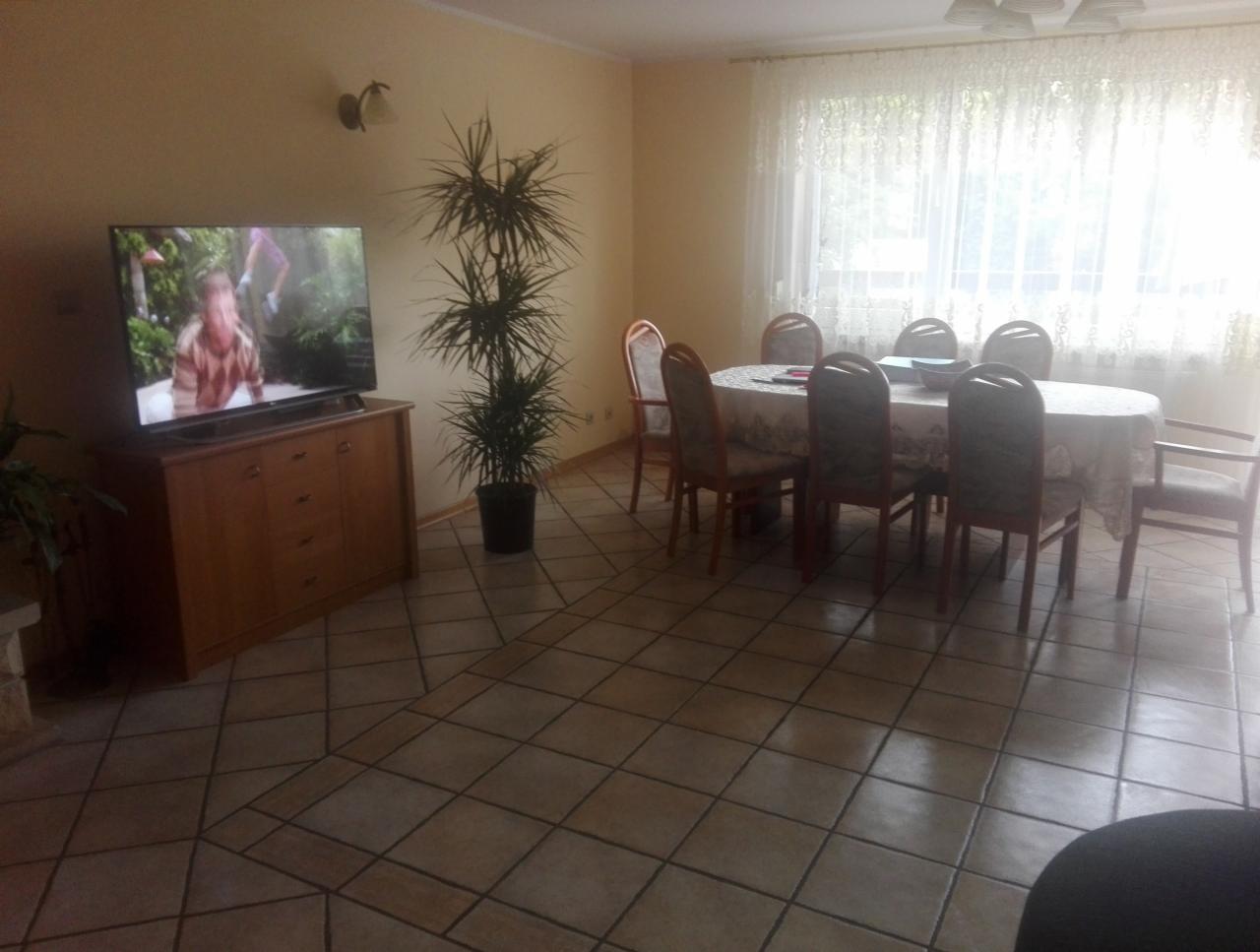 Jasny salon z parkietem w romby, stołem i krzesłami pod oknem z firankami, komodą z telewizorem i wysoką rośliną doniczkową.