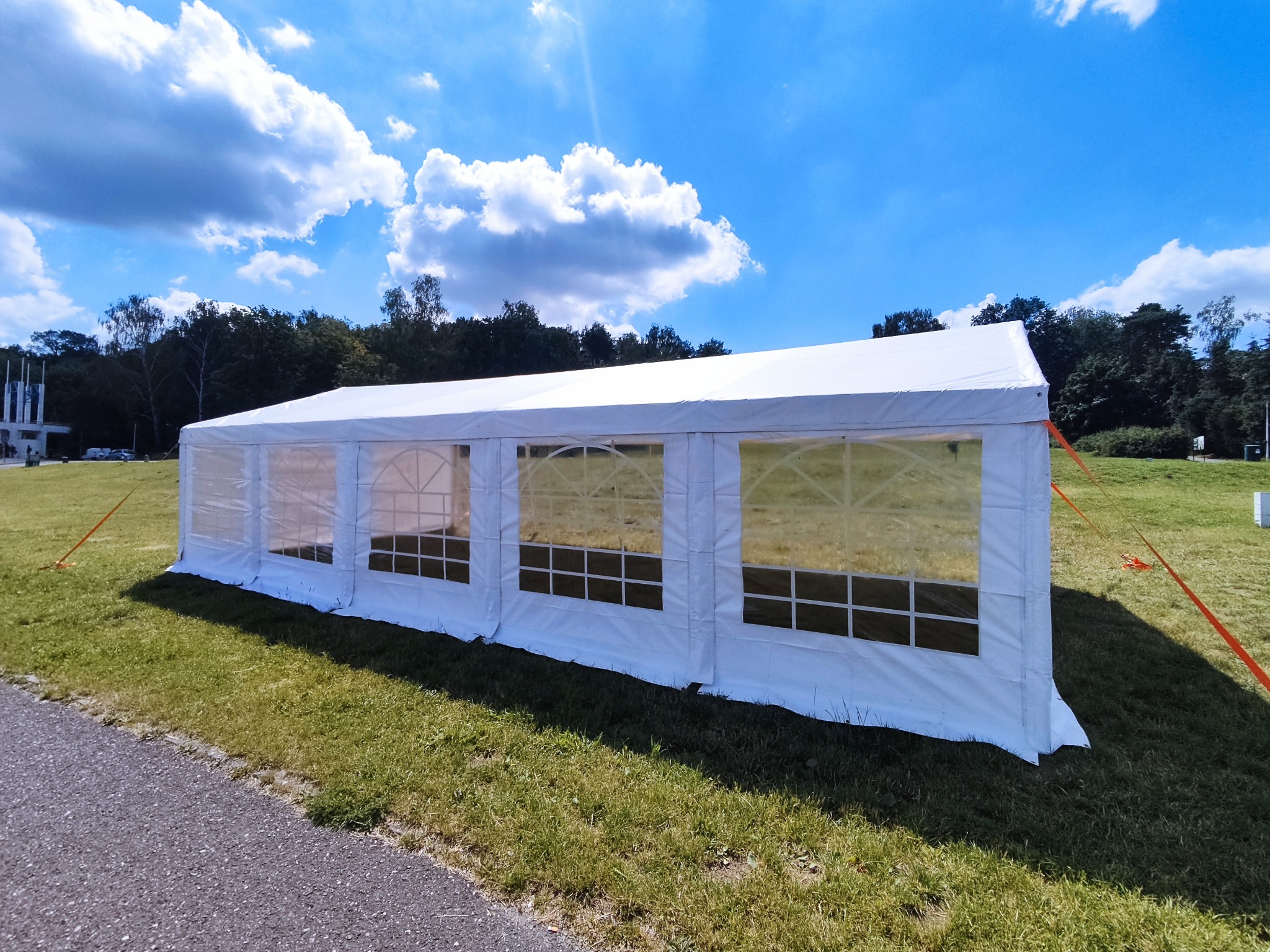 Biały namiot imprezowy 5x10 z przezroczystymi oknami na trawiastym terenie pod błękitnym niebem z chmurami, zabezpieczony pomarańczowymi linkami.