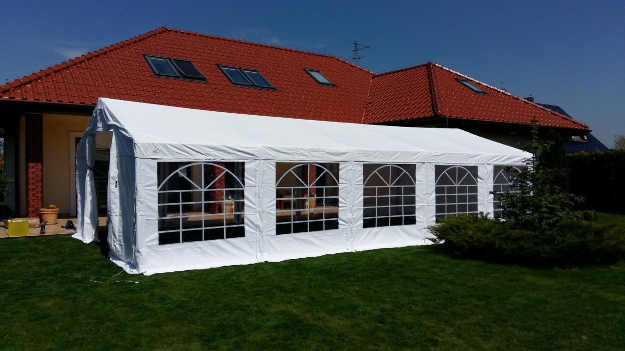 Biały namiot imprezowy z oknami w kształcie łuków, rozstawiony na trawniku przed domem z czerwoną dachówką i oknami dachowymi.