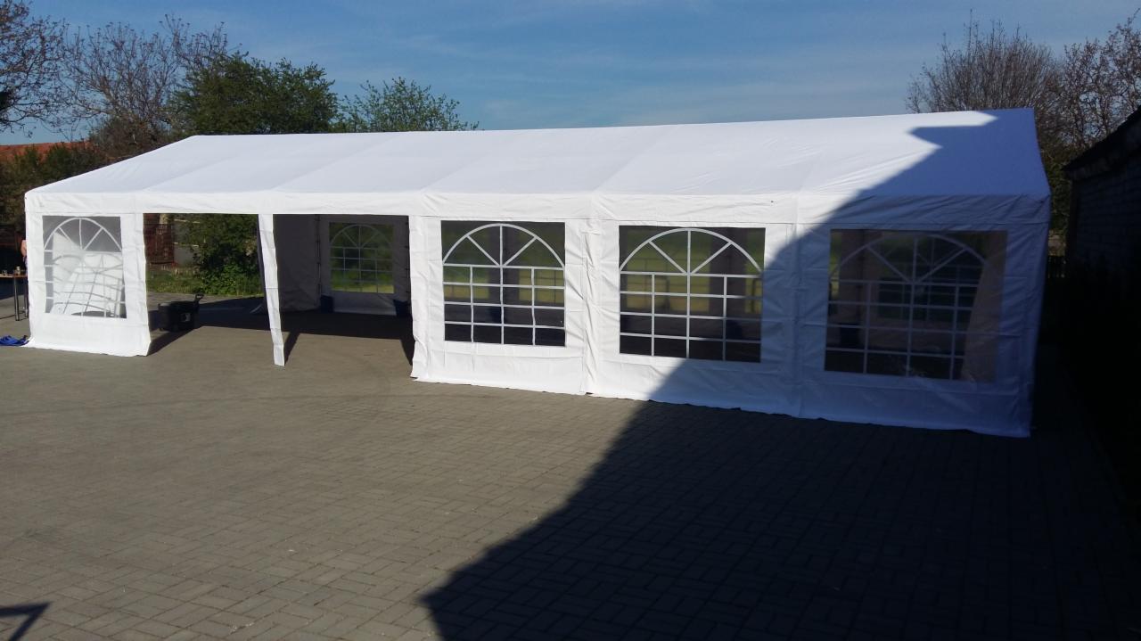 Rozłożony biały namiot plenerowy z oknami z białymi szprosami, postawiony na szarej kostce brukowej w słoneczny dzień.