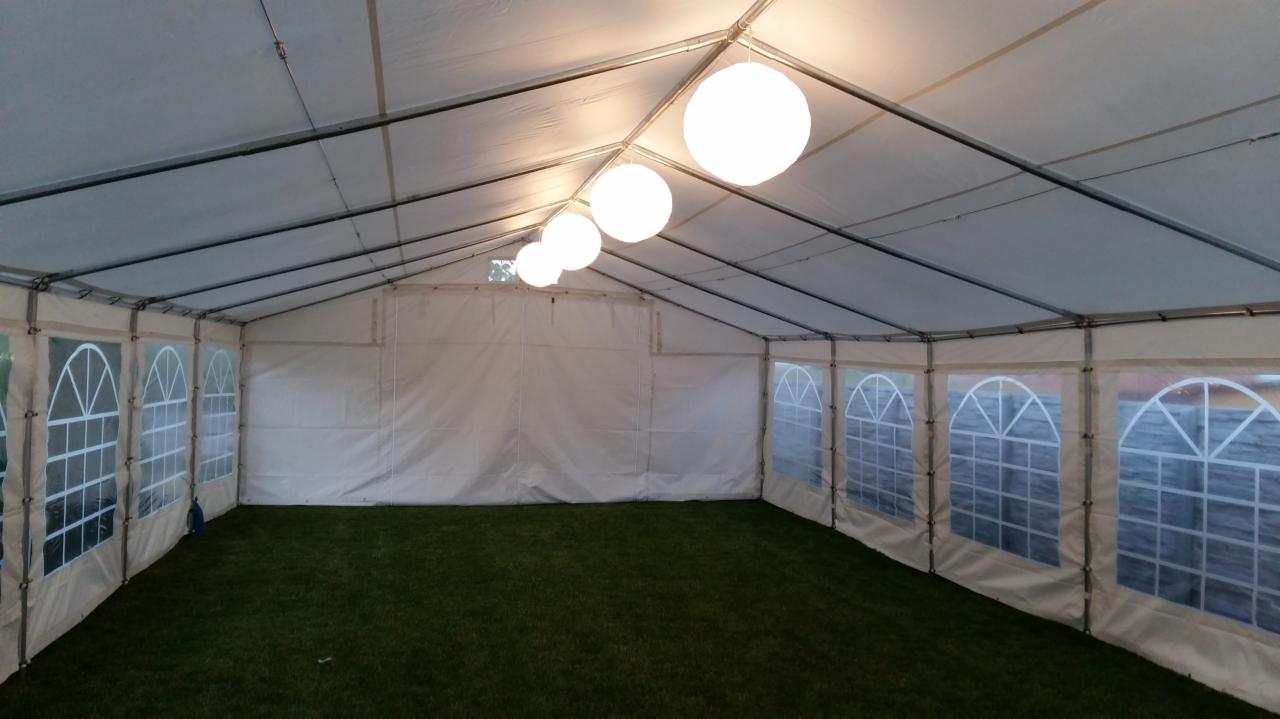 Wnętrze białego namiotu z metalową konstrukcją, oświetlonego wiszącymi kulami, z oknami po bokach i sztuczną trawą na podłodze.