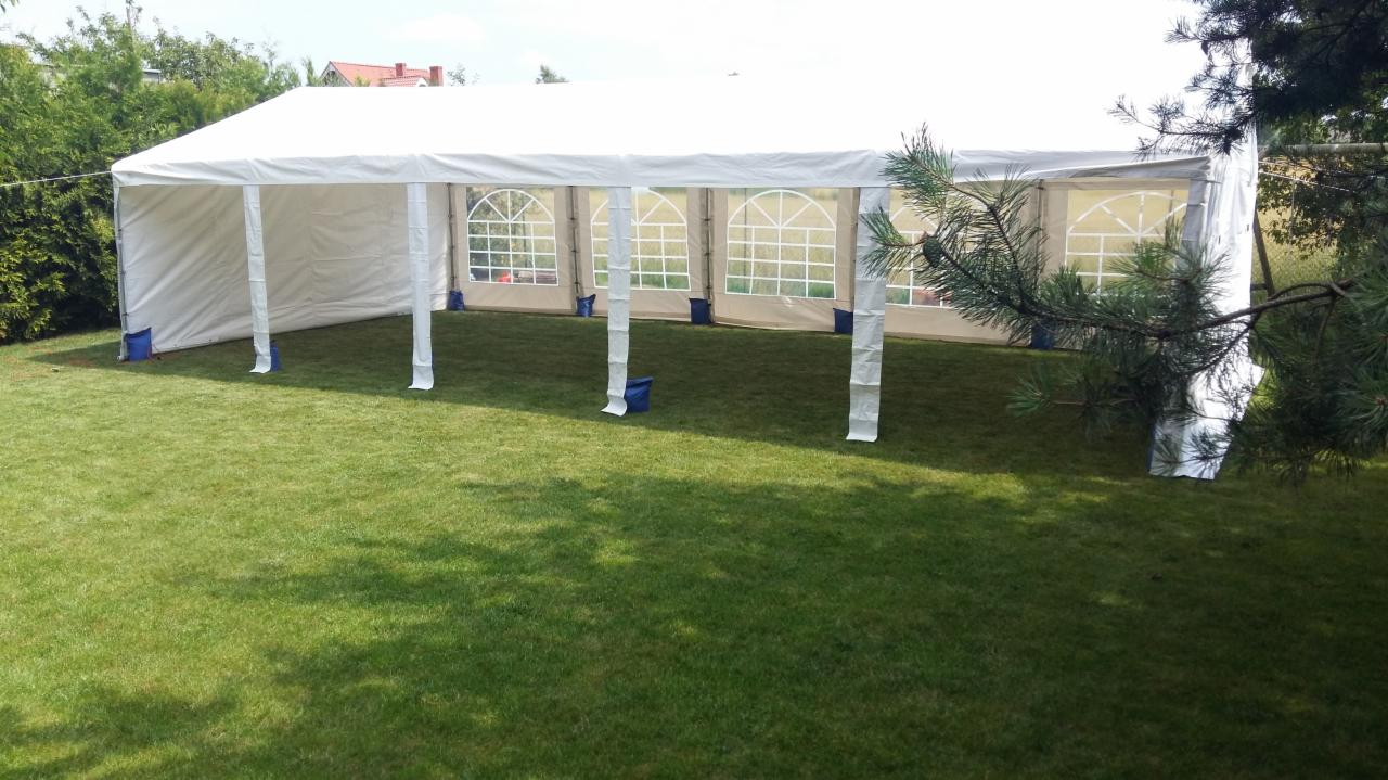 Rozłożony biały namiot plenerowy z oknami na trawniku, zabezpieczony niebieskimi obciążnikami, widok z boku przez gałąź drzewa.