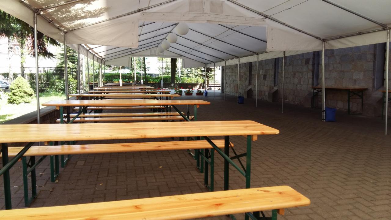 Rozstawiony biały namiot z rzędami drewnianych stołów i ławek na zewnątrz, przygotowany do imprezy plenerowej, widoczne drzewa w tle.