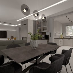 Modei Sztuka & Design - Wnętrze salonu z aneksem kuchennym w odcieniach szarości, z dużym stołem i krzesłami. Nad stołem wiszą trzy okrągłe lampy. W tle telewizor i zabudowa kuchenna.