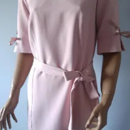 Różowa sukienka wizytowa z falbanką przy dekolcie i wiązanym paskiem, prezentowana na manekinie z widocznymi detalami takimi jak kokardki na rękawach i rozcięcia u dołu.