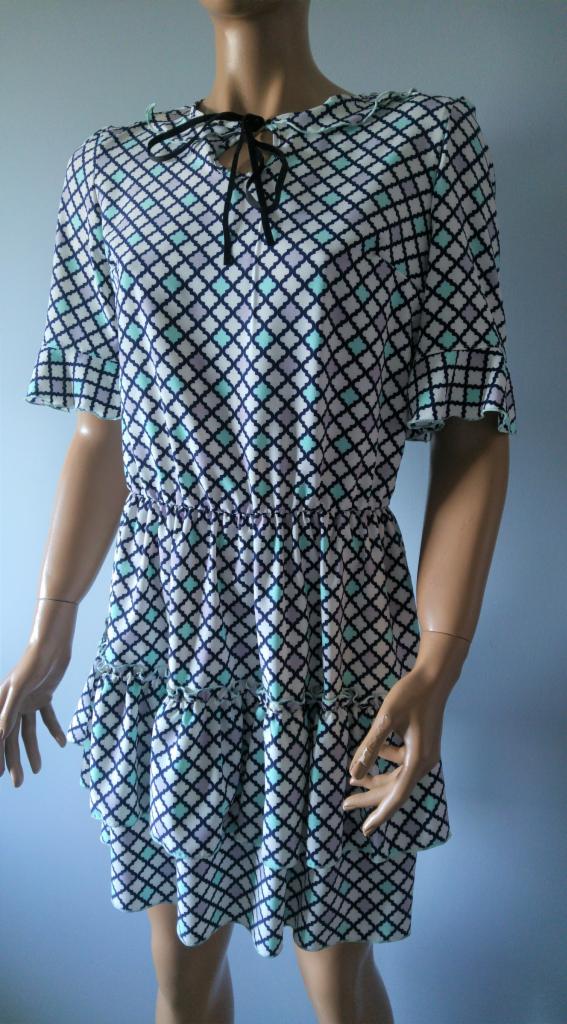 Krótka sukienka w biało-granatowo-miętowo-liliowy wzór karo, z falbankami na rękawach i dole, oraz czarną wstążką przy dekolcie, prezentowana na manekinie.