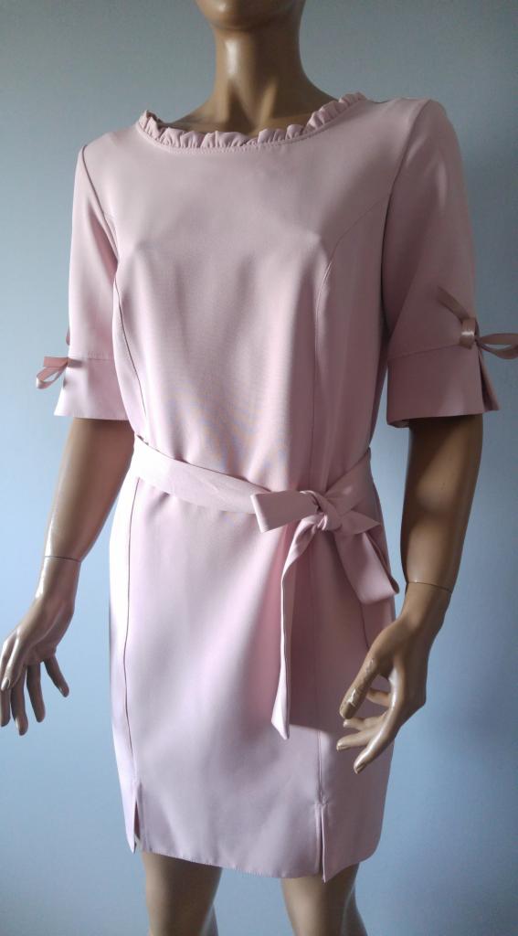 Różowa sukienka wizytowa z falbanką przy dekolcie i wiązanym paskiem, prezentowana na manekinie z widocznymi detalami takimi jak kokardki na rękawach i rozcięcia u dołu.