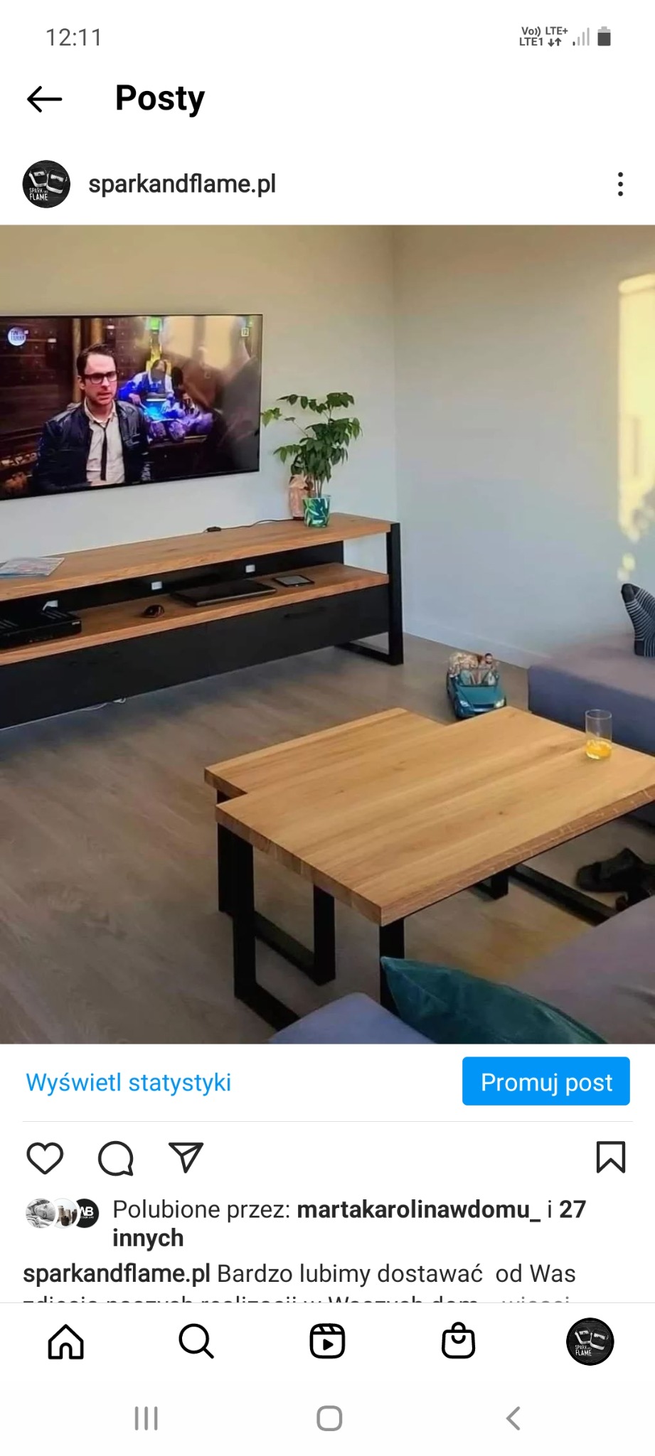 Stolik kawowy i szafka RTV z drewnianym blatem i czarną metalową podstawą w nowoczesnym salonie, widoczny telewizor i roślina doniczkowa.