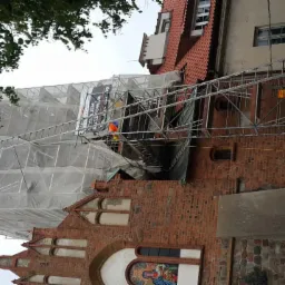 Budynek z czerwonej cegły, częściowo zakryty rusztowaniem i białą siatką ochronną, widoczny fragment mozaiki nad wejściem, fragment białego samochodu na pierwszym planie po prawej stronie.