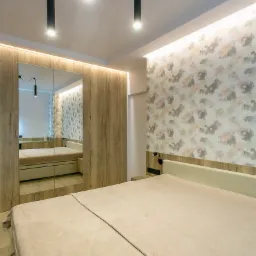 Nowoczesna sypialnia z łóżkiem w beżowej tonacji, szafą z lustrzanymi drzwiami i designerskimi lampami wiszącymi na tle ściany z delikatnym wzorem.