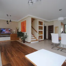 Jasne wnętrze domu jednorodzinnego z drewnianą podłogą, schodami z drewnianymi półkami na dekoracje, telewizorem i białymi krzesłami przy stole z kolorowym obrusem.