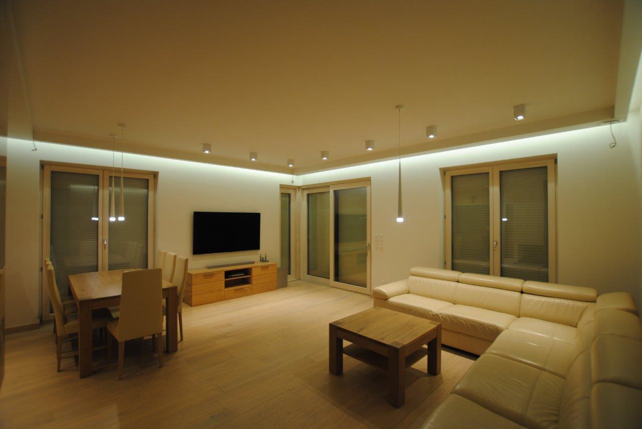 Jasne wnętrze salonu z jadalnią: drewniany stół z krzesłami, skórzana sofa narożna, szafka RTV i duży telewizor na ścianie, oświetlenie punktowe i wiszące lampy.