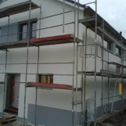 Dwupiętrowy dom w trakcie osuszania i wykończenia elewacji, widoczne rusztowania, częściowo pomalowana ściana na szaro na dole, okna i drzwi zabezpieczone folią.