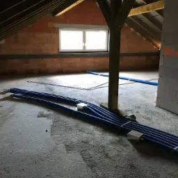 Poddasze w trakcie prac instalacyjnych: wiązka niebieskich rur wentylacyjnych leży na surowym betonowym podłożu, widoczne ceglane ściany i drewniana konstrukcja dachu.