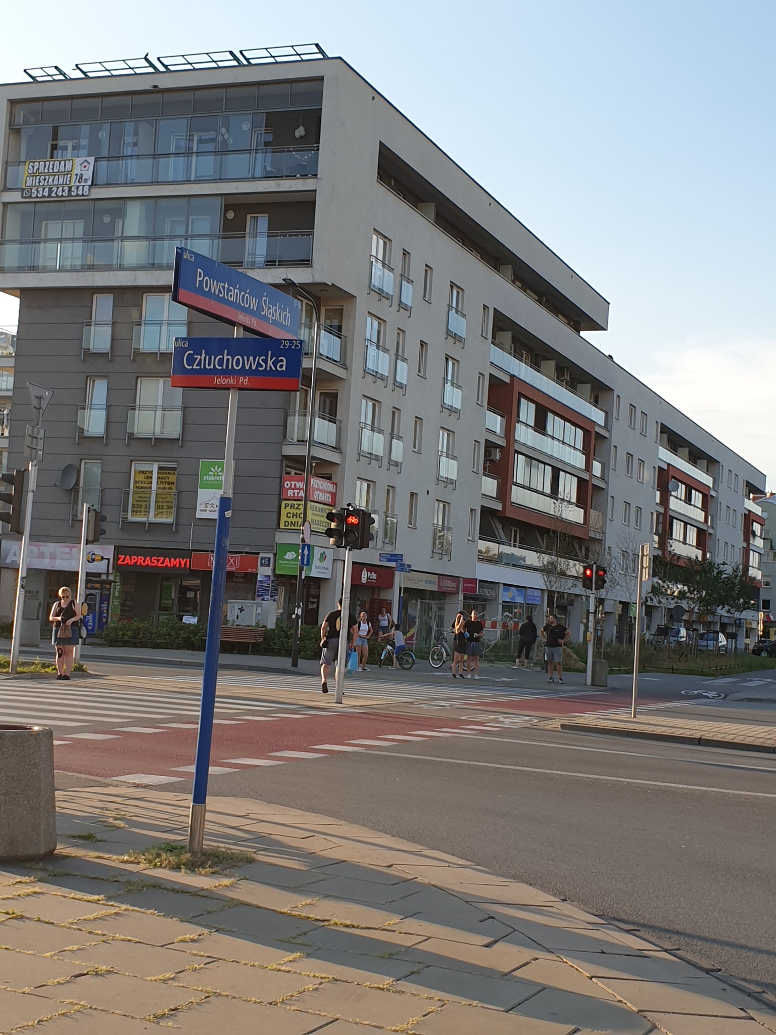 Ulica w dzielnicy Jelonki w Warszawie, z nowoczesnym budynkiem apartamentowym, znakami ulicznymi Powstańców Śląskich i Człuchowska, przejściem dla pieszych i grupą osób.