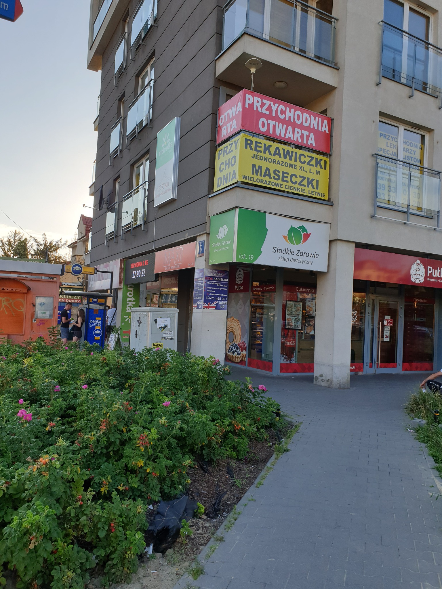 Budynek z różnorodnymi sklepami i przychodnią, widoczne szyldy 'Słodkie Zdrowie', 'Putka', 'Przychodnia Otwarta', bankomat oraz kwitnące krzewy przed budynkiem.