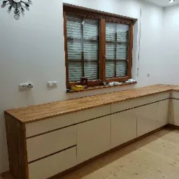 Długa szafka kuchenna z białymi frontami i drewnianym blatem, widok na okno z żaluzjami i zegar w kształcie sztućców na ścianie.