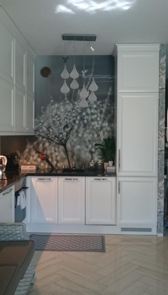 Nowoczesna kuchnia z białymi frontami szafek, czarnym blatem i dekoracyjną ścianą z motywem roślinnym, oświetlona wiszącą lampą z kloszami w kształcie kropli.