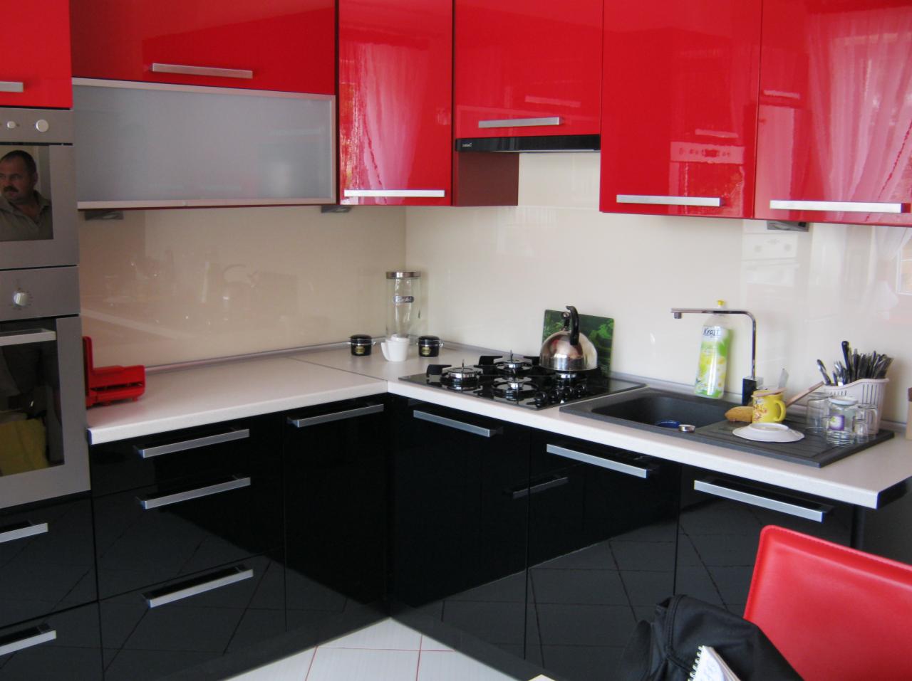 Nowoczesna kuchnia z czerwonymi szafkami górnymi i czarnymi dolnymi, biały blat i szklany panel nad blatem, widoczny fragment kuchenki gazowej i zlewu z naczyniami.