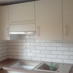 Kuchnia z białymi, prostokątnymi płytkami z dekoracyjnymi wstawkami, beżowe szafki wiszące i zlew z butelką płynu do naczyń.