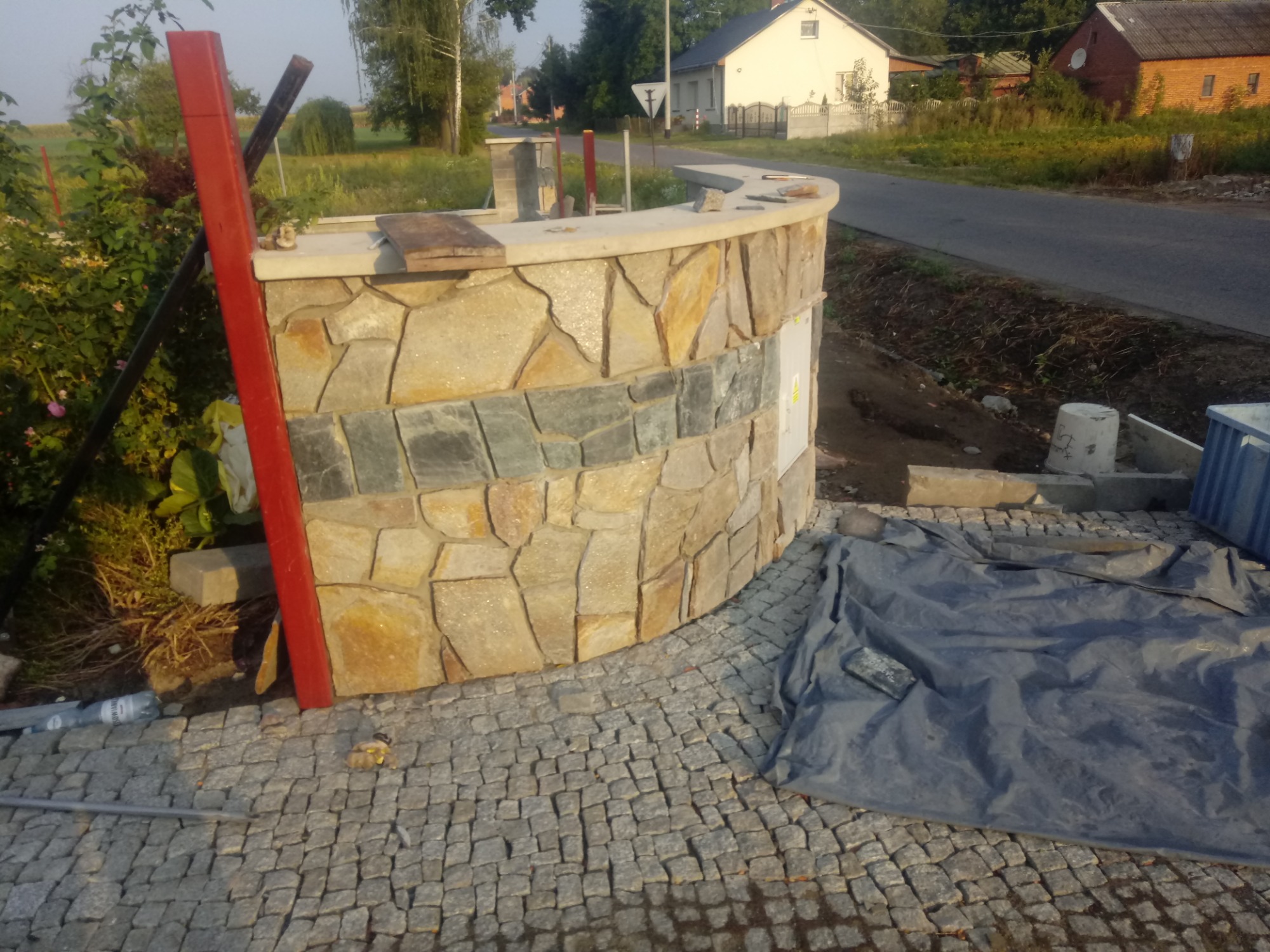 Nisko kątowe ujęcie półokrągłego murku z kamienia naturalnego w trakcie budowy, z widoczną kostką brukową i elementami rusztowania, na tle wiejskiej zabudowy.
