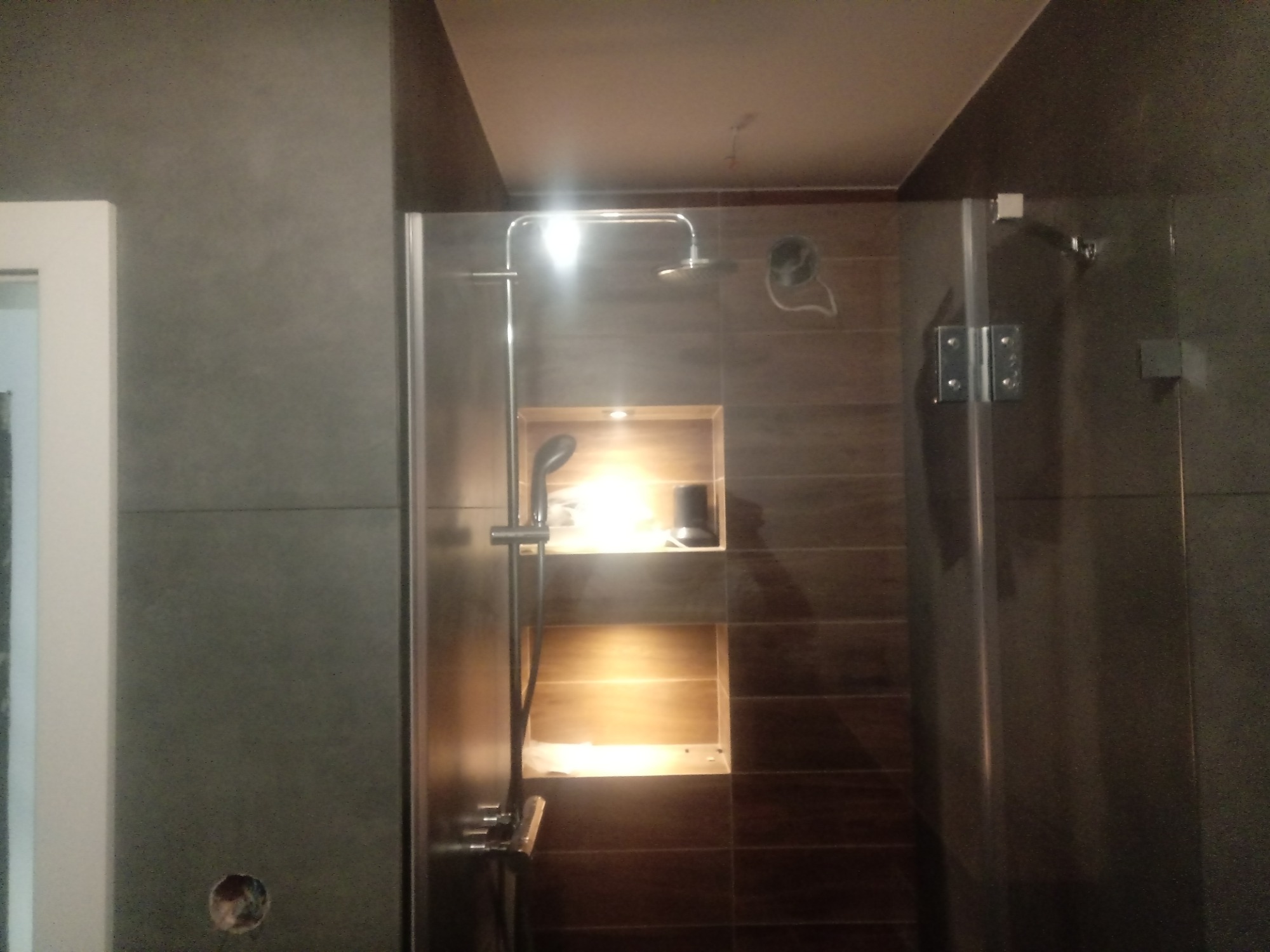 Kabina prysznicowa z deszczownicą i półkami w ścianie, wykończona ciemnymi płytkami imitującymi drewno.