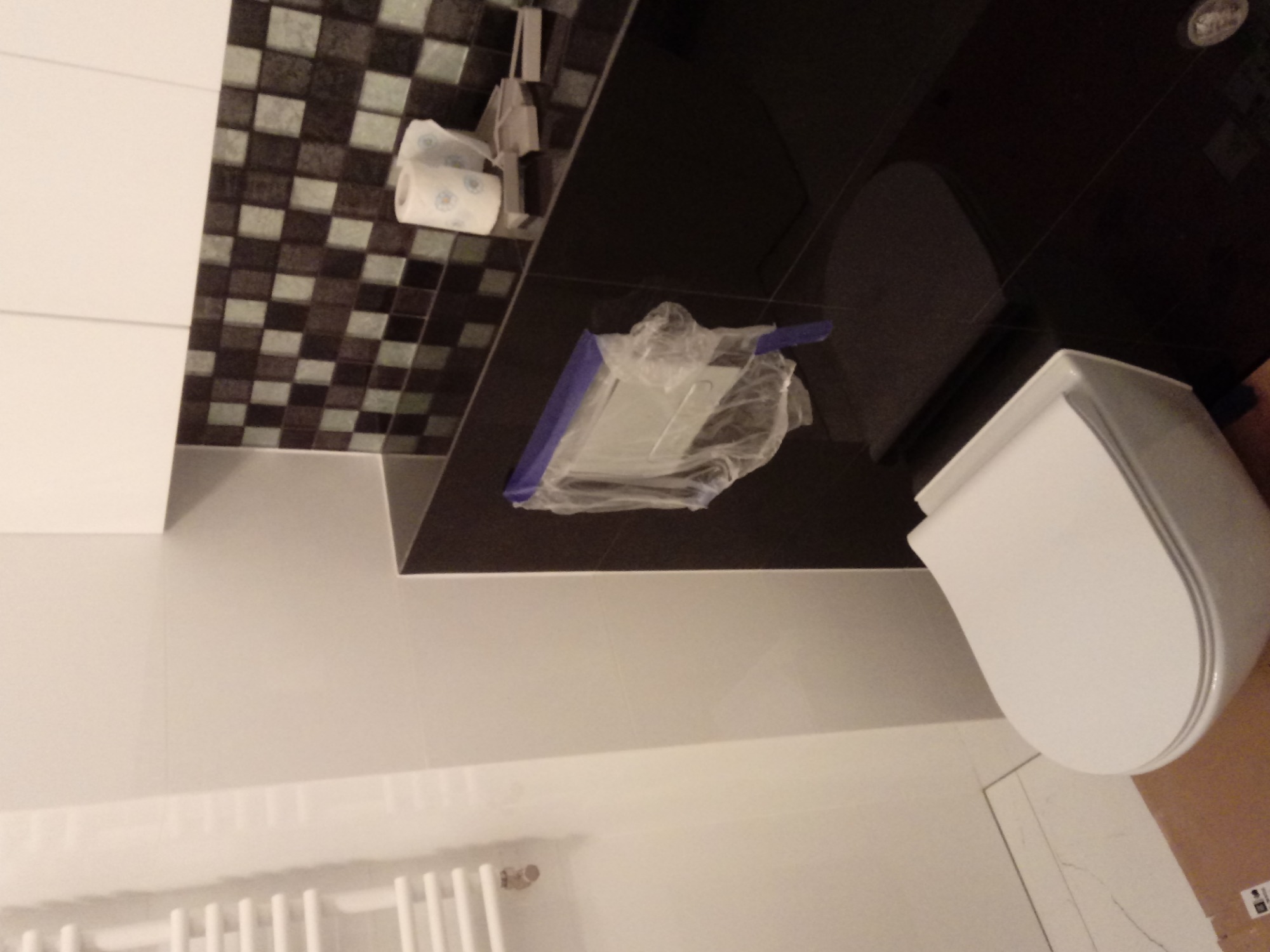 Nowa, biała toaleta w trakcie montażu w łazience z mozaikową ścianą, zabezpieczona folią ochronną, widoczny grzejnik i rolka papieru toaletowego.