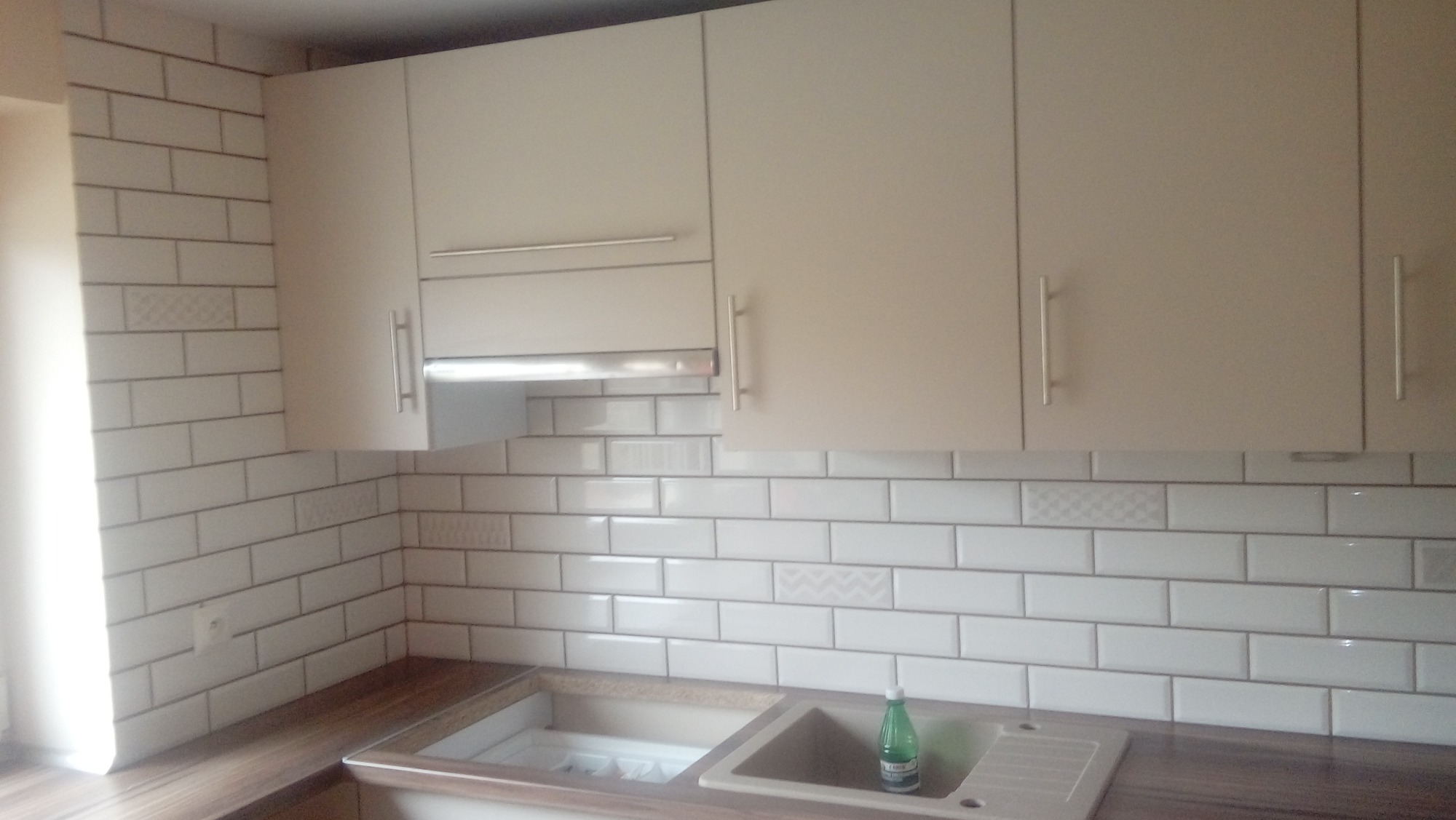 Kuchnia z białymi, prostokątnymi płytkami z dekoracyjnymi wstawkami, beżowe szafki wiszące i zlew z butelką płynu do naczyń.