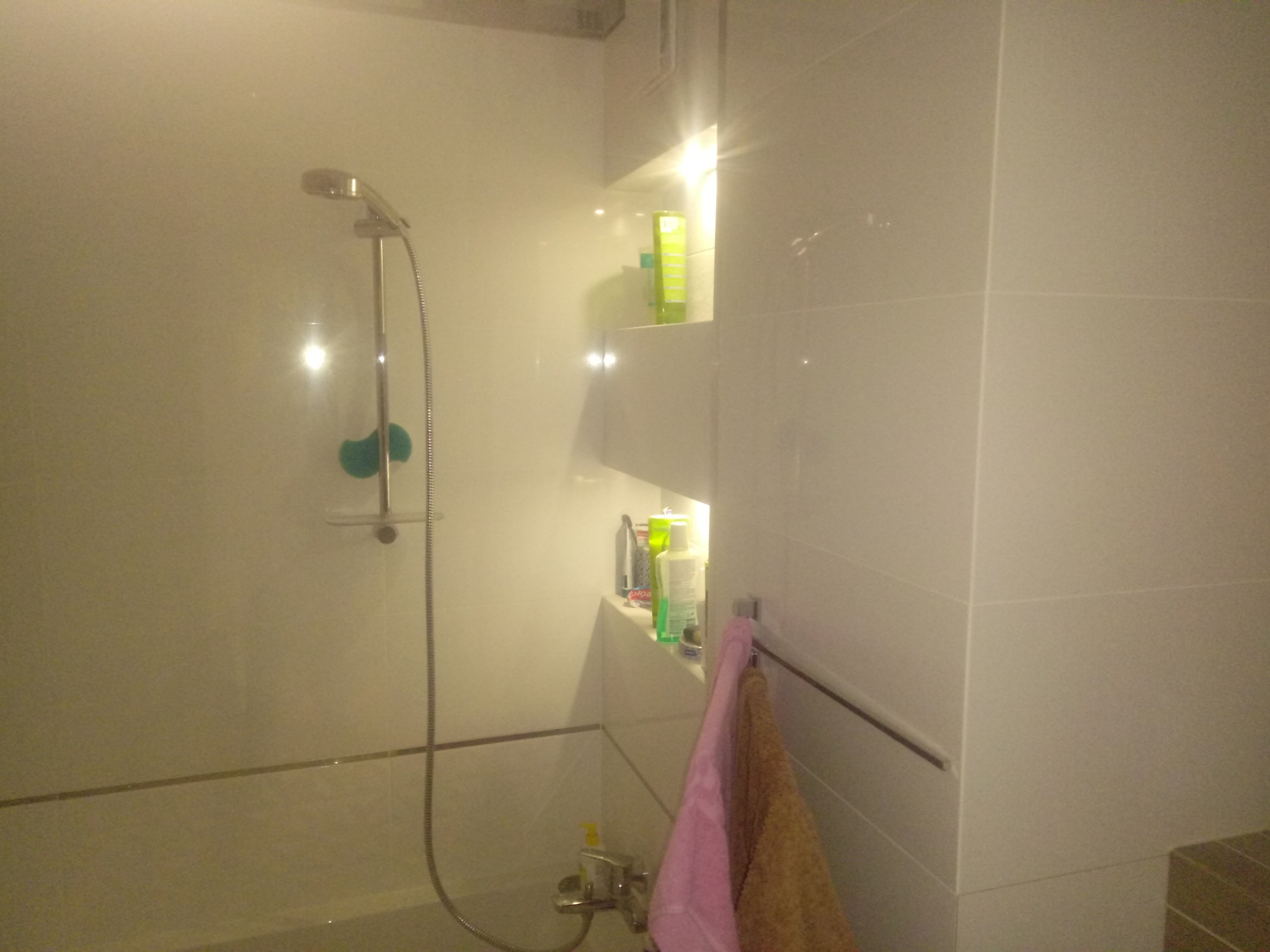 Biała łazienka z kabiną prysznicową, wbudowanymi półkami z kosmetykami i ręcznikami na relingu, widok z perspektywy osoby stojącej w wannie.