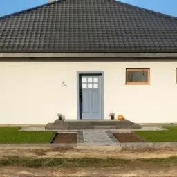 Nowoczesny, parterowy dom z szarą dachówką i jasną elewacją, drewnianymi żaluzjami w oknach i szarymi drzwiami wejściowymi, z widocznym samochodem dostawczym z tyłu, sugerujący prace wykończeniowe.