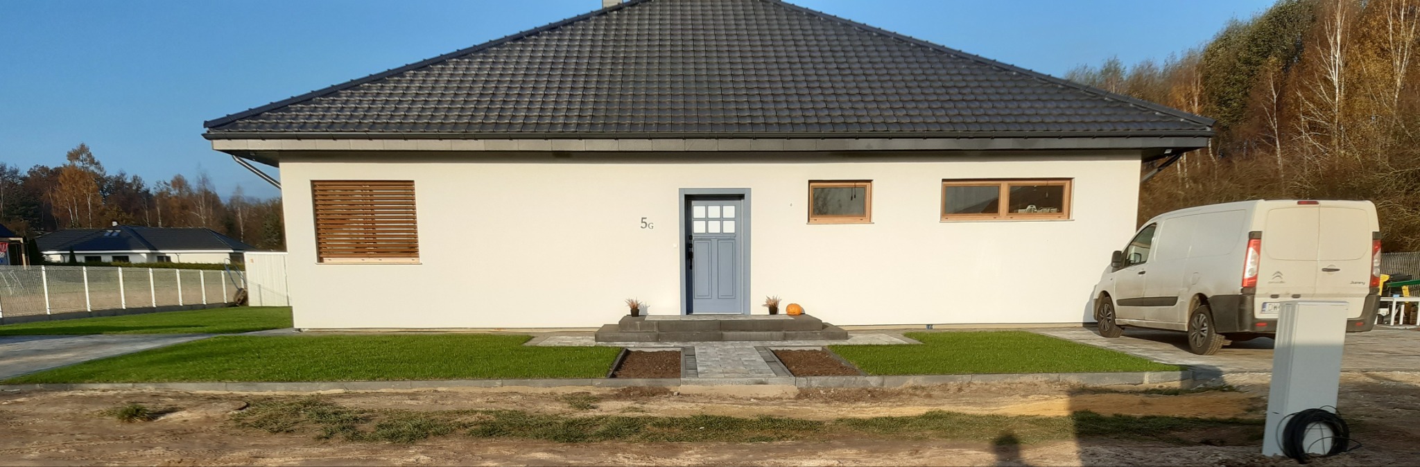 Nowoczesny, parterowy dom z szarą dachówką i jasną elewacją, drewnianymi żaluzjami w oknach i szarymi drzwiami wejściowymi, z widocznym samochodem dostawczym z tyłu, sugerujący prace wykończeniowe.