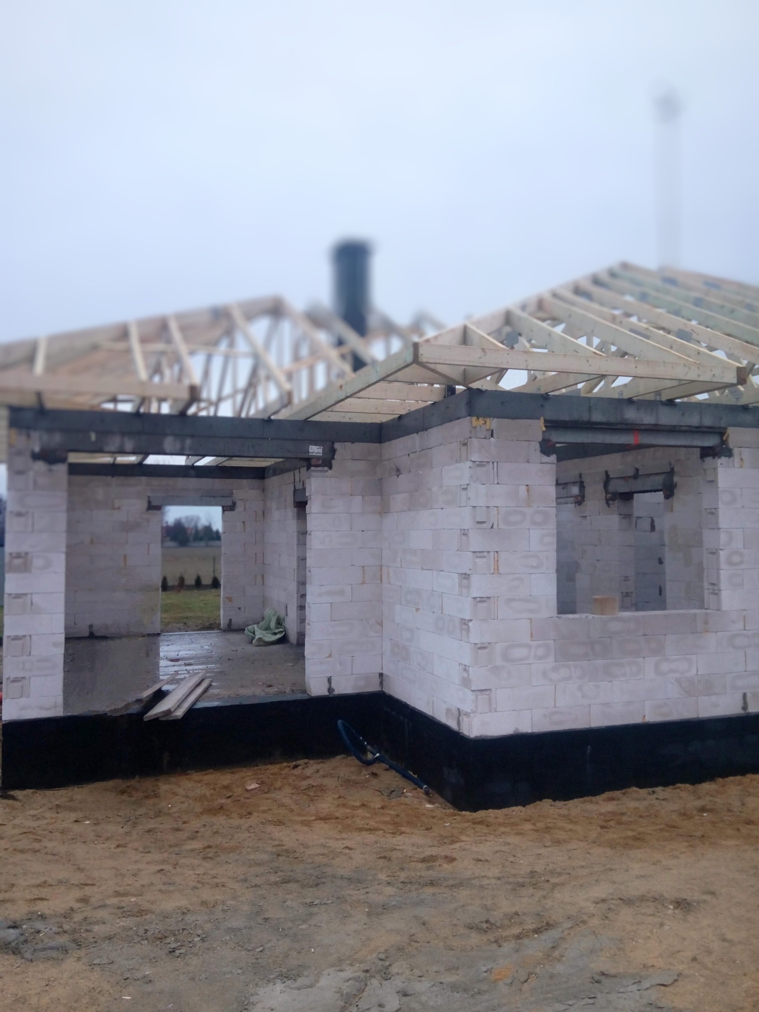 Surowy stan budynku jednorodzinnego z białych bloczków, widoczne ściany bez ocieplenia, czarna izolacja fundamentów, drewniana konstrukcja dachu w trakcie budowy, teren wokół budynku piaszczysty.