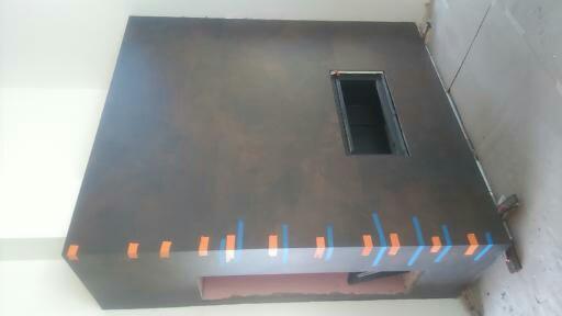 Ściana z otworem, pokryta ciemnym tynkiem dekoracyjnym z pomarańczowo-niebieskimi paskami taśmy malarskiej w dolnej części, widoczne ślady po pracach wykończeniowych.