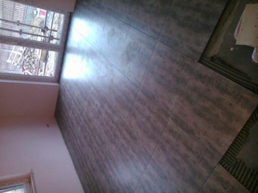 Układanie ciemnych, podłużnych płytek podłogowych w pomieszczeniu, widoczne niedokończone fragmenty i okno z widokiem na zewnątrz.