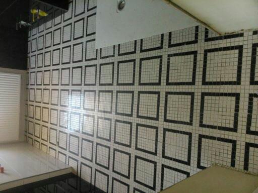 Ściana wyłożona białymi mozaikami z czarnymi obramowaniami tworzącymi wzór kwadratów, widoczne fragmenty pomieszczenia z żaluzjami i czarnymi elementami wystroju.