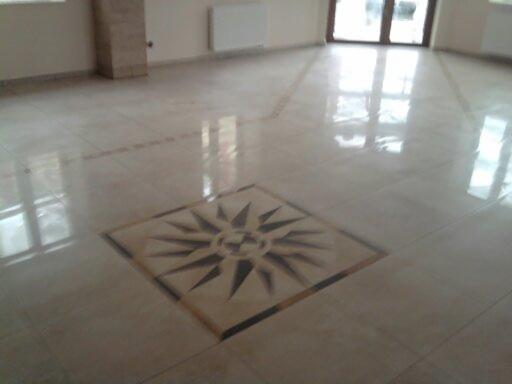 Jasna, polerowana podłoga z płytek z dekoracyjnym motywem słońca w centrum, odbijająca światło okien.