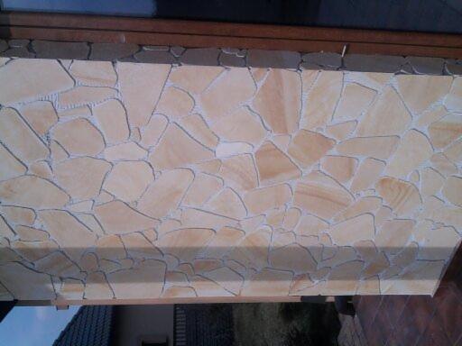 Ściana zewnętrzna wykończona nieregularnymi, beżowymi płytkami kamiennymi o zróżnicowanej fakturze, ułożonymi z widoczną fugą. Widoczne elementy dachu i fragment budynku w tle.