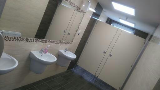 Publiczna toaleta z trzema umywalkami, lustrem z dekoracyjną obwódką i kabinami WC z jasnymi drzwiami.