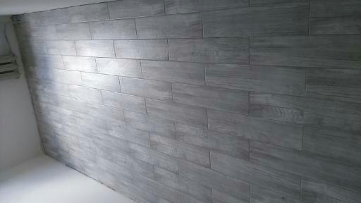 Ściana wyłożona szarymi, podłużnymi płytkami ceramicznymi imitującymi drewno, ułożonymi poziomo, widoczne fugi i fragment białej ściany z grzejnikiem po lewej stronie.
