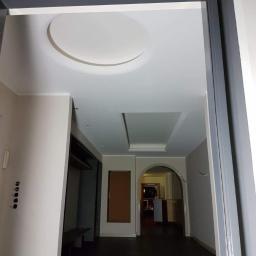 MEGA BUD Arkadiusz Zakrzewski - Wnętrze mieszkania po remoncie, widok na sufit z okrągłym i prostokątnym podwieszanym elementem, szara rama drzwi z żółtą masą szpachlową w górnym rogu.