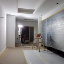 MEGA BUD Arkadiusz Zakrzewski - Wnętrze w trakcie malowania: ściany przygotowane do malowania, zabezpieczona folią podłoga i ściana, drabina z wiadrem, oświetlenie budowlane na statywie.