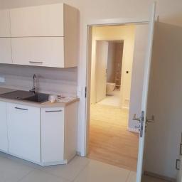 MEGA BUD Arkadiusz Zakrzewski - Nowoczesna, jasna kuchnia z białymi szafkami i szarą mozaiką nad blatem, widok na łazienkę z toaletą i drabinką przez otwarte drzwi.