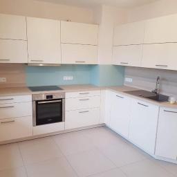 MEGA BUD Arkadiusz Zakrzewski - Nowoczesna, jasna kuchnia z białymi szafkami, drewnianym blatem i turkusowym panelem szklanym nad płytą grzewczą. Widoczne białe płytki na podłodze i minimalistyczne uchwyty szafek.