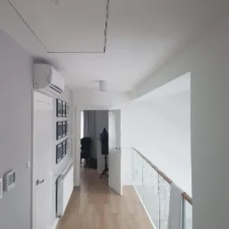 Nowoczesny korytarz na poddaszu z drewnopodobnymi panelami, szarą ścianą z dekoracjami, klimatyzacją, grzejnikiem i szklaną balustradą z drewnianym poręczą.