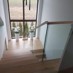 MEGA BUD Arkadiusz Zakrzewski - Drewniane schody z przeszkloną balustradą w nowoczesnym domu, widok z góry na dół, duże okno z widokiem na ogród, dekoracyjne rośliny w donicach.