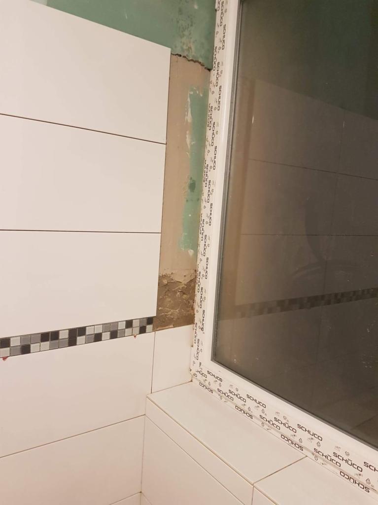 Białe płytki ścienne z dekoracyjnym paskiem mozaiki w łazience, widoczne surowe wykończenie ściany obok okna z ramą PCV.