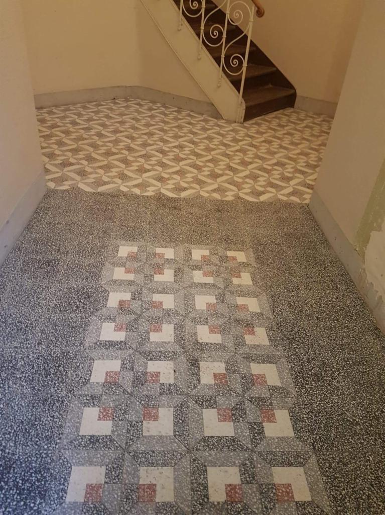 Wzorzysta podłoga z płytek w przedpokoju, z widocznymi schodami z dekoracyjną, białą balustradą w tle, wykonana z dwóch rodzajów płytek: mozaiki i kwadratowych płytek z ornamentem.